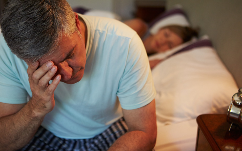 A man sitting on his bed, struggling to sleep due to his bad back