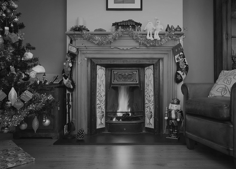 A black and white image of a fireplace decorated with tinsel and stockings next to a Christmas tree in a living room.