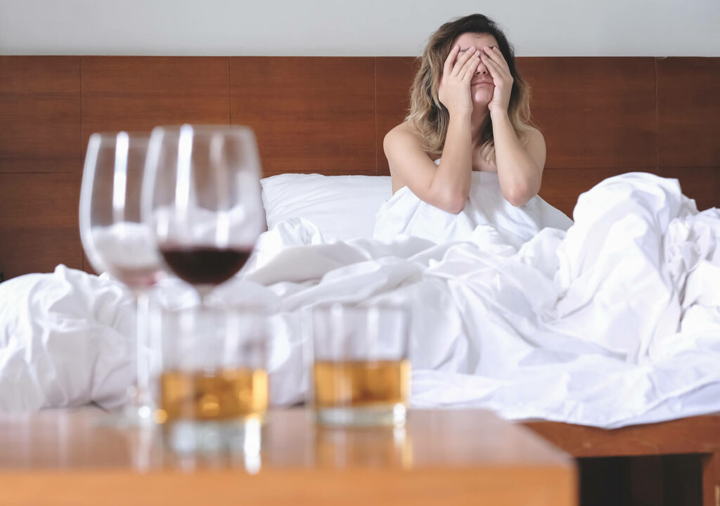 A woman sitting on a wooden bed with her hands covering her face with glasses of wine and whiskey on the bedside table.