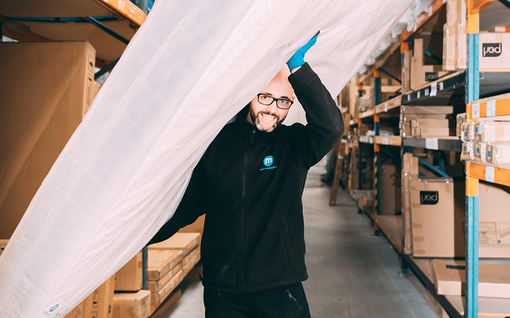 A Mattress Online warehouse staff member smiling while carrying a mattress through the warehouse.