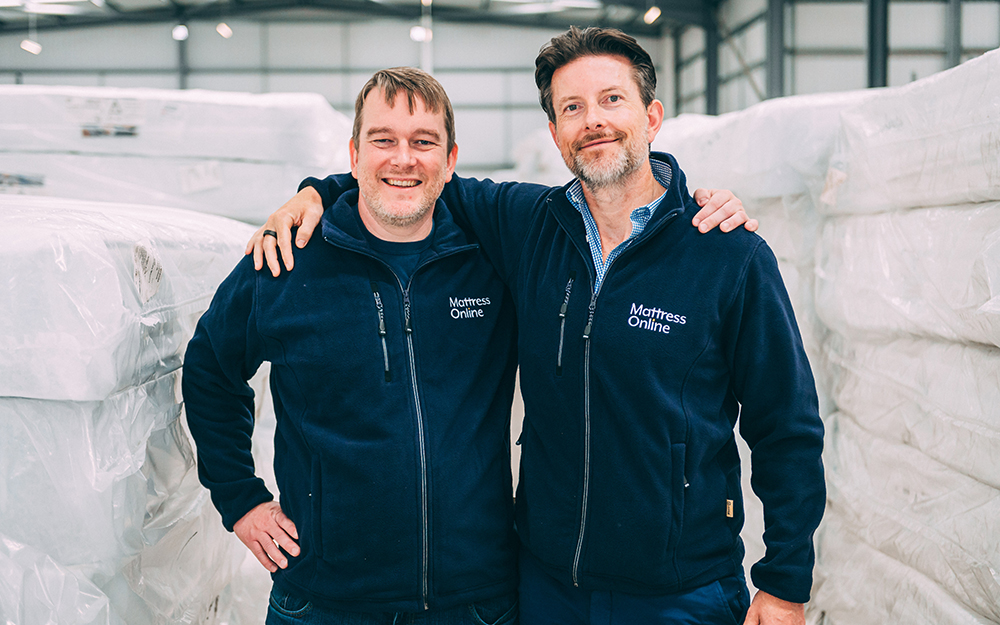 Martin and Steve Adams smiling together in a Mattress Online warehouse, arms around each other.