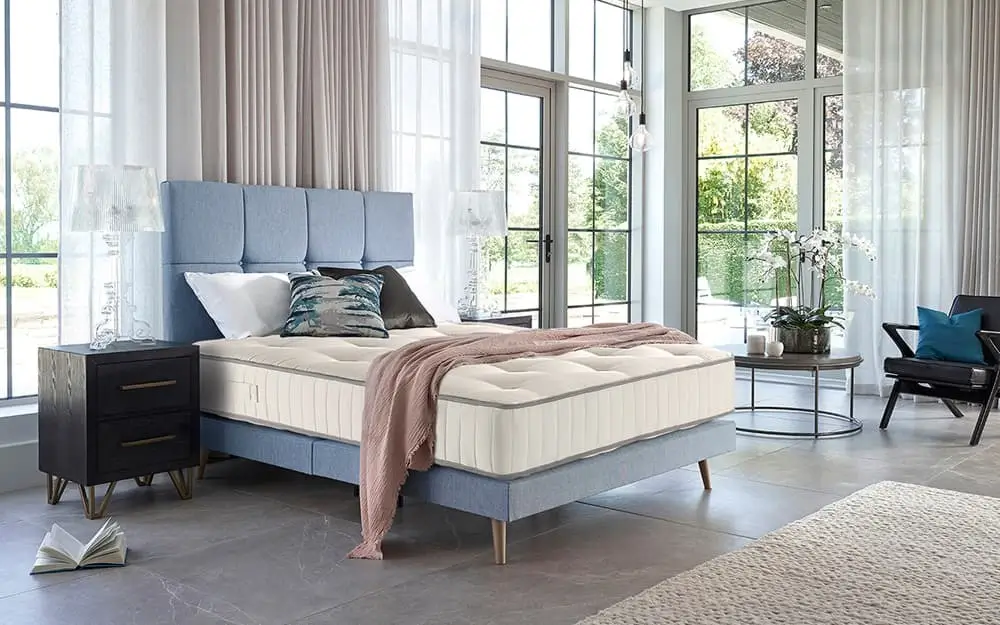 The Harrison Spinks Luxury Essential 750 Pocket Mattress on a blue fabric bed frame in an open bedroom with floor to cieling windows, overlooking a large greenn garden space.